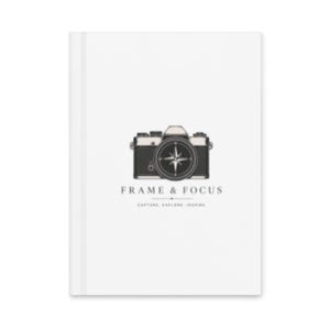 Può includere: Un taccuino bianco con una grafica di fotocamera in bianco e nero e il testo "FRAME & FOCUS". La grafica della fotocamera ha un design a rosa dei venti al centro. Il taccuino è vuoto.