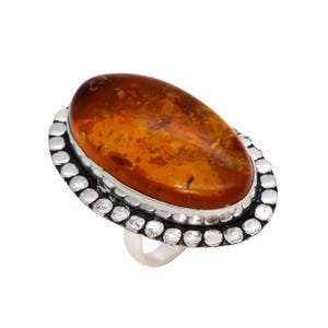 Op de afbeelding: Ovale barnsteen edelsteenring met een zilveren band. De barnsteen steen heeft een warme, doorschijnende oranje-bruine kleur, gezet in een zilveren bezel met een gestippelde rand. De band van de ring is van gepolijst zilver.