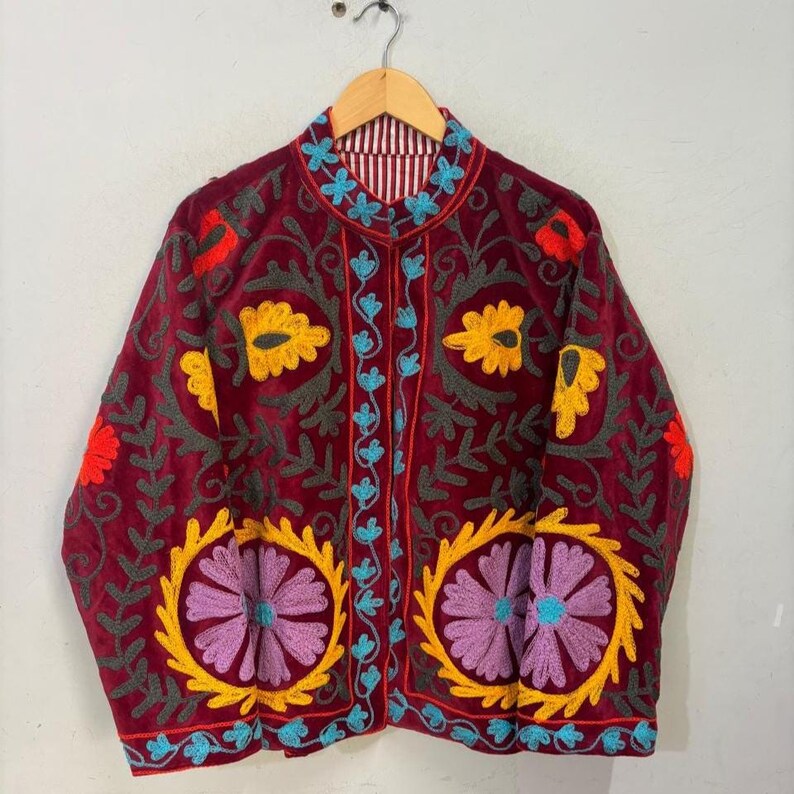 May include: A burgundy velvet jacket with intricate floral embroidery in yellow, purple, red, and teal. The jacket features a mandarin collar and a striped lining. The embroidery creates a striking contrast against the dark background, showcasing a bohemian style.