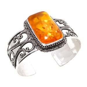 Op de afbeelding: Een zilveren manchetarmband met een grote, rechthoekige barnsteen. De barnsteen heeft een warme, doorschijnende oranje kleur. De armband heeft een sierlijk, open ontwerp met krulpatronen.