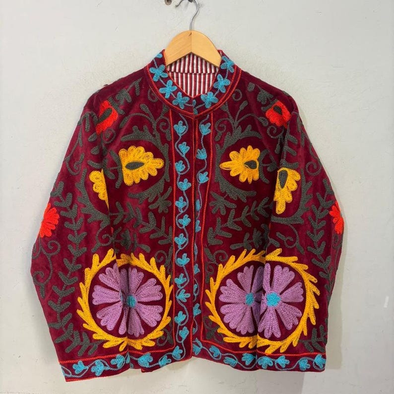 May include: A burgundy velvet jacket with intricate floral embroidery in yellow, purple, red, and teal. The jacket features a mandarin collar and is lined with a striped fabric. The design includes large floral motifs and detailed vine patterns.