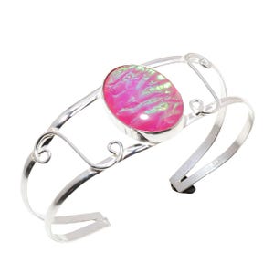 Puede incluir: Una pulsera de plata con un óvalo de piedra preciosa rosa iridiscente. La pulsera tiene un diseño delicado y abierto con remolinos y curvas decorativas. La piedra preciosa es el punto focal.