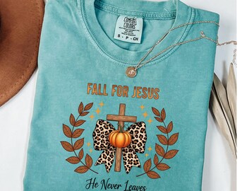 May include: A turquoise Comfort Colors t-shirt featuring the text "FALL FOR JESUS" above a cross, pumpkin, and leopard print bow design. The phrase "He Never Leaves" is below the design. A gold necklace is draped across the shirt.