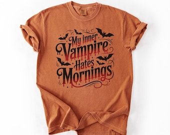 Op de afbeelding: Een roestoranje t-shirt met korte mouwen en een zwart-rode gradiënt grafische print met de tekst My inner Vampire Hates Mornings, omringd door kleine zwarte vleermuissilhouetten en decoratieve krullende lijnen op de borst.