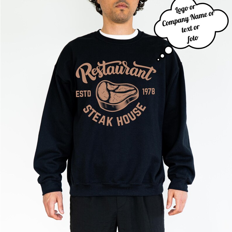 Może przedstawiać: Czarna bluza z białym okrągłym dekoltem. Z przodu brązowa grafika z napisem "Restaurant Steak House" i rokiem "1978". Projekt zawiera ilustrację steku.
