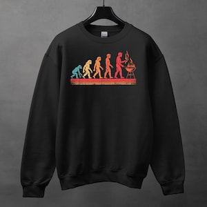 May include: Black sweatshirt featuring a retro-style graphic of human evolution ending with a person grilling. The graphic uses a color palette of blue, yellow, orange, and red. The design is printed on the front of the sweatshirt.