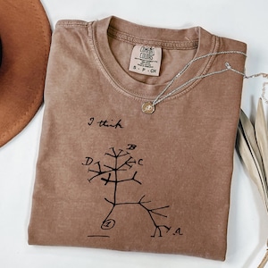 Puede incluir: Una camiseta marrón con el texto "I think" y un diagrama negro en forma de árbol. Un collar plateado con un pequeño colgante descansa sobre la camiseta. La camiseta está etiquetada como "Comfort Colors" y es talla pequeña.