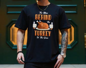 Może przedstawiać: Czarny t-shirt z napisem "The Man Behind The Turkey In The Oven". Projekt zawiera pieczonego indyka i ilustracje kciuków w górę w kolorze pomarańczowym i białym. Koszulka ma krótkie rękawy.