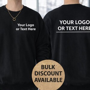 Może przedstawiać: Czarna bluza z napisem "Your Logo or Text Here" z przodu i z tyłu. Z przodu widoczny jest złoty naszyjnik. Okrągły znak z napisem "Bulk Discount Available" znajduje się na dole. Bluza jest w jednolitym czarnym kolorze.