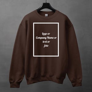 Może przedstawiać: Brązowa bluza z białym prostokątnym wzorem pośrodku. Tekst wewnątrz prostokąta brzmi "Logo lub Nazwa firmy lub tekst lub foto". Bluza wisi na czarnym wieszaku.