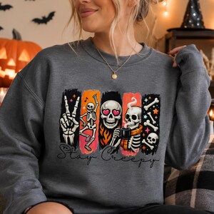 May include: A dark gray crewneck sweatshirt featuring a graphic print of skeletons, flames, and stars. The design includes a skeleton making a peace sign, a dancing skeleton, a skull with heart eyes, and a skeleton drinking coffee. The text reads Stay Creepy.