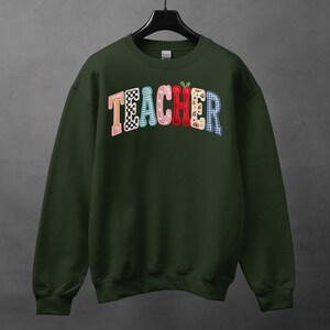 May include: A dark green sweatshirt featuring the word "TEACHER" in a vibrant, multi-patterned design. The letters include checkerboard, floral, and plaid patterns. A small apple graphic is above the letter "E".