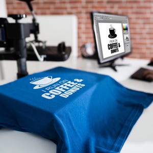 Może przedstawiać: Niebieska koszulka z białym napisem "I RUN ON COFFEE & DONUTS". Obraz przedstawia koszulkę na białej powierzchni, z monitorem komputera wyświetlającym ten sam wz&oacute;r. W tle widoczna jest prasa termotransferowa.