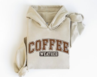 Puede incluir: Sudadera con capucha beige con las palabras "COFFEE WEATHER" en letras marrones. La sudadera con capucha tiene una capucha con cordón y un bolsillo de canguro. Las mangas están enrolladas.