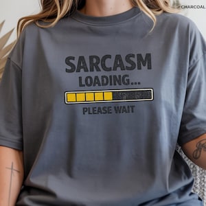 May include: Charcoal gray t-shirt with the text "SARCASM LOADING... PLEASE WAIT" in black. The design includes a yellow and black loading bar graphic, creating a humorous and relatable statement.