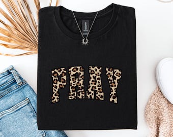 May include: Black t-shirt with the word "PRAY" in a leopard print design. A silver necklace with a sunburst pendant rests on the shirt. The shirt is folded and placed on a white surface with blue jeans and a white shoe.