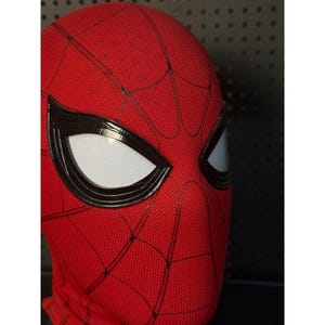 May include: A red Spider-Man mask with black webbing and large white eye lenses. The mask is made of a textured fabric and has a black border around the eyes. The mask is a close-up shot.