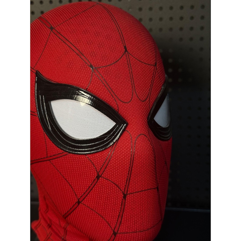 May include: A red Spider-Man mask with black webbing and large white eye lenses. The mask is made of a textured fabric and has a black border around the eyes. The mask is a close-up shot.