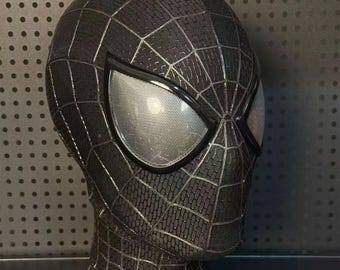 May include: A black Spider-Man mask with a textured surface and webbed design. The mask features large, mesh-covered eye openings with a silver reflective appearance. The mask is displayed on a black bust.