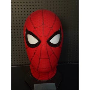 May include: A red Spider-Man mask with black webbing and large white eye lenses. The mask is displayed on a black stand, showcasing the character's iconic design. The mask is made of a textured fabric.