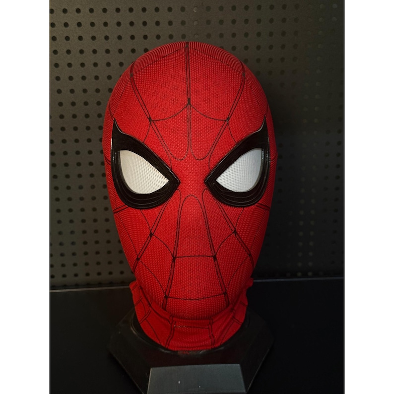 May include: A red Spider-Man mask with black webbing and large white eye lenses. The mask is displayed on a black stand, showcasing the character's iconic design. The mask is made of a textured fabric.