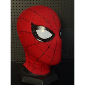 May include: A red and black Spider-Man mask with white eye lenses. The mask has a textured surface and a web pattern design. The mask is displayed on a black stand, showcasing the superhero's iconic look.