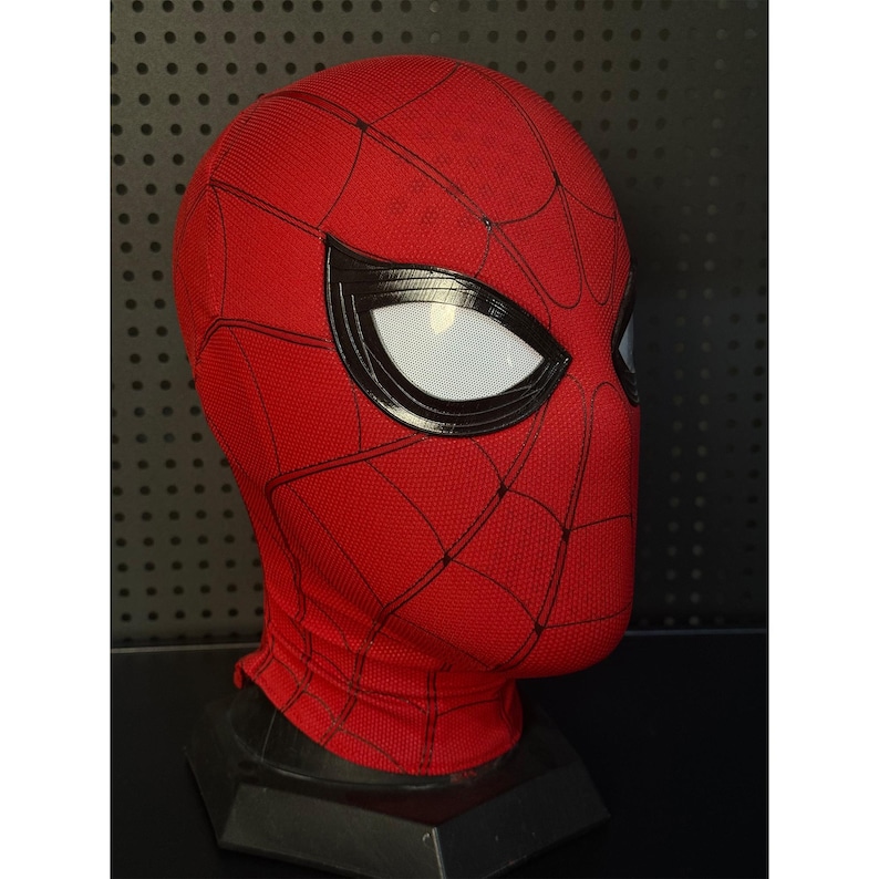May include: A red and black Spider-Man mask with white eye lenses. The mask has a textured surface and a web pattern design. The mask is displayed on a black stand, showcasing the superhero's iconic look.