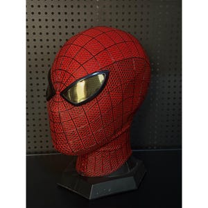 May include: A red and black Spider-Man mask with gold-tinted eye lenses. The mask has a textured surface with a web pattern and is displayed on a black stand. The mask is a replica of the superhero's headwear.