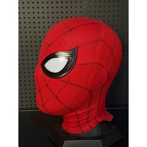 May include: A red Spider-Man mask with a black web pattern and large white eye lenses outlined in black. The mask is made of a textured fabric and is displayed on a black stand.