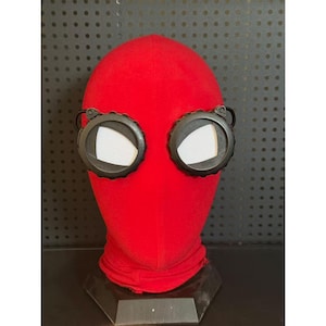 May include: A red fabric hood with black-rimmed goggles. The goggles have white mesh lenses. The hood is displayed on a black stand against a dark background.
