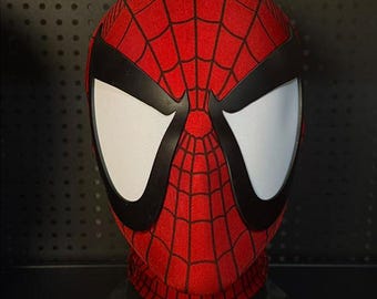 May include: A red and black Spider-Man mask with a web pattern. The mask has large white eye lenses outlined in black. The mask is made of a textured material and is displayed on a black stand.