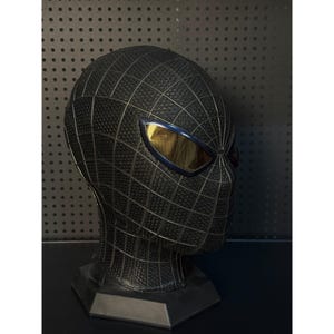 May include: A black Spider-Man mask with a textured surface and a webbed pattern. The eye lenses are gold with a blue border. The mask is displayed on a black stand.