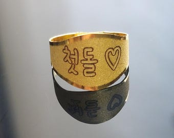 Puede incluir: Un anillo dorado con los caracteres coreanos "첫돌" y un símbolo de corazón grabado. El anillo se refleja en una superficie oscura.