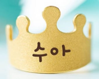 Puede incluir: Un anillo con forma de corona dorada con los caracteres coreanos "수아" impresos. El anillo tiene una forma de corona clásica con cinco puntas. El fondo es una mezcla borrosa de azul y blanco.