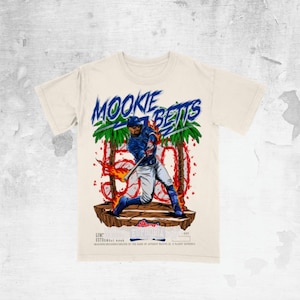 May include: Cream-colored t-shirt featuring a baseball player graphic with the text "MOOKIE BETTS" in blue and green. The design includes palm trees and flames, with the number "500" prominently displayed. The shirt has short sleeves.