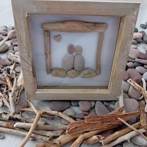 May include: A framed pebble art piece depicting two figures and a heart, made from natural stones and driftwood. The artwork is set within a light-coloured wooden frame, creating a rustic aesthetic. The frame measures approximately 20 cm by 20 cm.