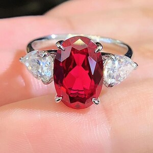 May include: A silver ring featuring a large, oval-cut, vibrant red gemstone as the centerpiece, flanked by two pear-shaped, clear gemstones. The ring is set on a hand, showcasing the jewelry's design and color.