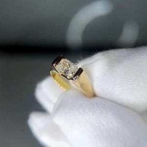 Könnte beinhalten: Ein goldener Ring mit einem rechteckigen, klaren Edelstein, der in einer dunklen Lünette gefasst ist. Das polierte Goldband kontrastiert mit dem klaren Edelstein. Der Ring wird vor einem weißen Stoff gehalten. Der Edelstein scheint ein Diamant zu sein.