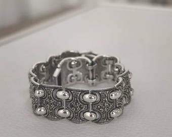 May include: A silver-toned bracelet with an ornate design. The bracelet features a series of oval and circular elements, with a textured pattern. The clasp is visible, and the bracelet is displayed on a white surface. Jewelry.