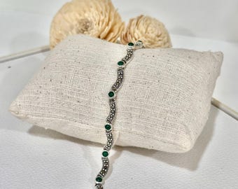 May include: A silver bracelet with a wavy design, featuring small, round, green gemstones and decorative silver accents. The bracelet is displayed on a small, square, off-white cushion.