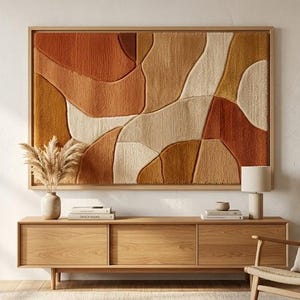 May include: A large, rectangular wall hanging with an abstract design in shades of orange, beige, and brown. The artwork is framed in light wood and hangs above a wooden console table. The room is decorated in neutral tones.