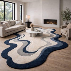 May include: An area rug with an irregular, organic shape featuring layered, wavy bands of colour in deep navy blue, slate grey, and cream. The rug has a thick, tufted texture and is positioned on a light-coloured wooden floor in a living room.