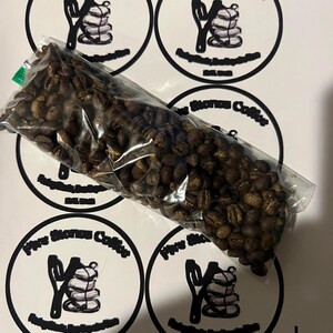 May include: A clear plastic bag filled with roasted coffee beans, resting on a white surface with multiple circular stickers. The stickers feature a black and white logo with the text "Five Stones Coffee".