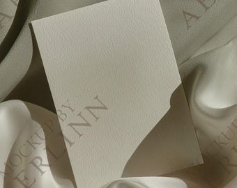 Puede incluir: Una tarjeta rectangular de color blanquecino con textura descansa sobre una tela fluida de color claro. La tarjeta está en ángulo, con sombras que crean profundidad. Las palabras "MOCKUP BY AERLINN" son débilmente visibles.
