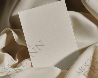 Puede incluir: Una tarjeta rectangular en blanco, de color blanquecino, con una superficie texturizada, apoyada sobre una tela crema drapeada. La tela tiene pliegues y sombras suaves, creando una sensación de profundidad. La tarjeta está colocada en ángulo, sugiriendo una maqueta o presentación.