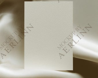 Puede incluir: Una tarjeta rectangular en blanco, de color blanquecino, con una superficie texturizada, sobre un fondo de tela fluida de color crema. La tela tiene pliegues y reflejos suaves. Las palabras "MOCKUP BY AERLINN" están impresas en la tela.