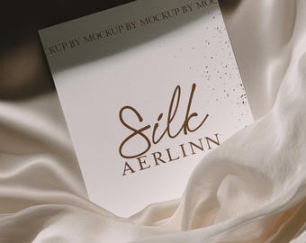 Puede incluir: Una tarjeta blanca con la palabra "Silk" en fuente marrón y el nombre "AERLINN" debajo. La tarjeta está sobre un fondo de tela de seda fluida de color crema. La parte superior de la tarjeta tiene el texto "MOCKUP BY MOCKUP BY MOC".