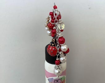 May include: A decorative charm with red beads, white pearls, and a ladybug pendant. The charm is attached to a silver chain and hangs over a bottle with the text "art glitter".