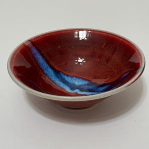 May include: A small, handmade ceramic bowl with a glossy finish. The bowl features a deep red exterior with a vibrant blue streak across the interior. The rim is a light cream color. This decorative bowl is ideal for serving small portions.