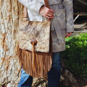 May include: A brown and white cowhide leather crossbody bag with a flap closure and long fringe. The bag has a brown leather strap and a wooden button closure. The bag is worn over the shoulder.